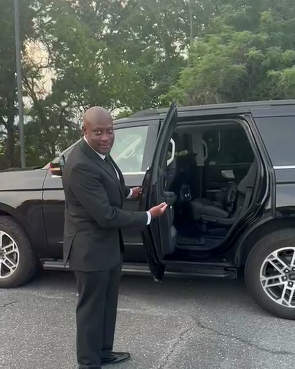 Chauffeur in a dark suit holding the rear passenger door of a black SUV open