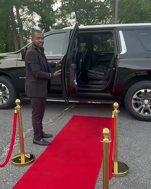 Chauffeur in a dark suit standing at the open rear door of a black Chevrolet Suburban at the end of a red carpet