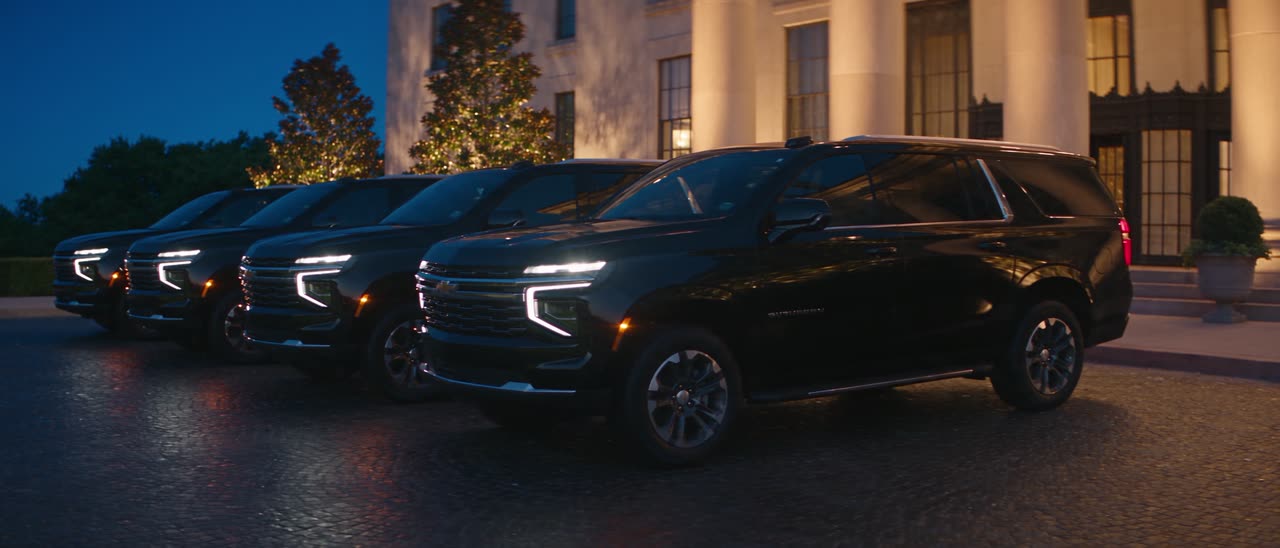 A line of black Chevrolet Suburbans on the forecourt of a lit hotel entrance at blue hour